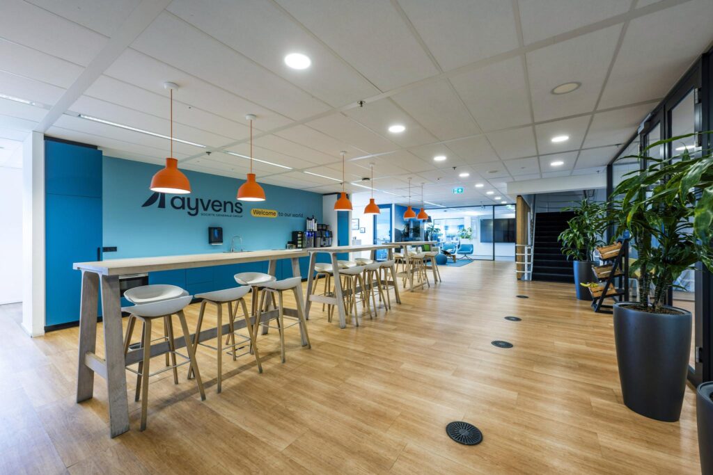 Moderne bedrijfskeuken en ontmoetingsruimte bij Ayvens aan de Johan Huizingalaan, met hoge tafels, barkrukken en oranje hanglampen.